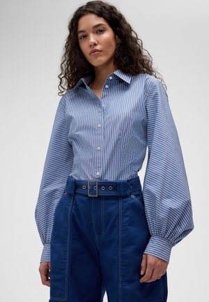 Femme aux cheveux bouclés portant une chemise à rayures bleues et blanches à boutons avec des manches ballon et un pantalon bleu marine taille haute avec une ceinture.