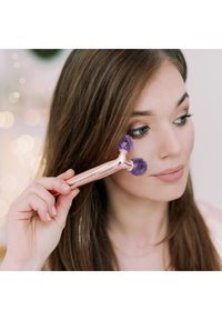 Różowo-złoty wałek do twarzy z dwoma zaokrąglonymi końcówkami z fioletowego kamienia trzymany przy policzku; użytkownik ma długie włosy i neutralny makijaż, miękkie tło.