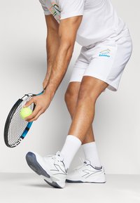 Hvide atletiske shorts og t-shirt med multicolor logoer. Personen holder et tennisracket med en gul bold, iført hvide sneakers og sokker.