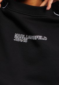 Černá mikina s žebrovaným límcem, na přední straně s bílým výšivkovým textem "KARL LAGERFELD JEANS". Text je tučný a má moderní design.