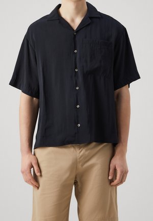 Mand iført en sort kortærmet skjorte med brystlomme og beige shorts, står mod en ensfarvet baggrund.