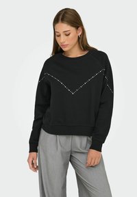 Jeune femme portant un sweat à capuche noir avec des clous argentés en motif chevron et un pantalon large gris clair, se tenant devant un fond blanc.