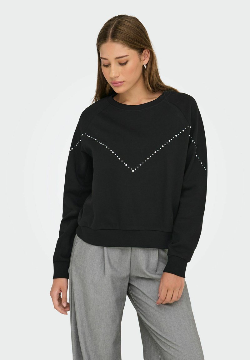 Jeune femme portant un sweat à capuche noir avec des clous argentés en motif chevron et un pantalon large gris clair, se tenant devant un fond blanc.
