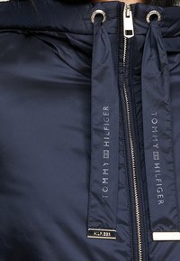 Veste bleu marine avec une fermeture éclair dorée et deux cordons de serrage, arborant un logo "Tommy Hilfiger" en texte blanc.