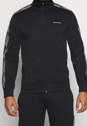 Osoba ubrana w czarny dres z logo Jack & Jones na piersi i rękawach, stojąca na jasnym, jednolitym tle.