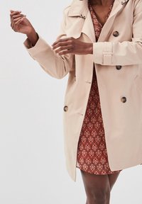 Persoon die de mouwen van een beige trenchcoat aanpast, gedragen over een rood patroonjurk, terwijl hij tegen een eenvoudige lichte achtergrond staat.