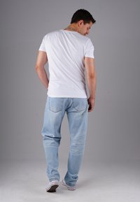 Man standing with back turned, wearing a white t-shirt, light blue jeans, and white sneakers against a plain gray background.
