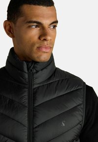 Čierna prešívaná vesta s vysokým golierom, vzorom v tvare šípky, zipsom a nenápadným logom na hrudi. Ľahký materiál.