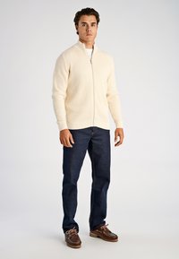 Kremowy sweter na suwak o teksturze diamentów, zestawiony z ciemnoniebieskimi dżinsami i brązowymi butami na sznurowadła z fakturowaną podeszwą.