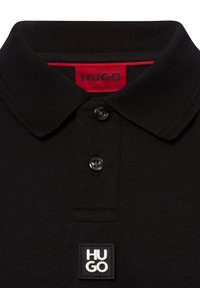 Camiseta tipo polo negra de tela suave con cuello clásico, dos botones y una etiqueta cuadrada con el logo "HUGO" en blanco en el dobladillo. Etiqueta roja en el interior.