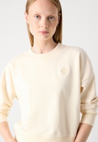 Sweatshirt crème avec des poignets et une taille côtelés, présentant un petit logo en diamant en relief sur la poitrine. Tissu doux et lisse avec une coupe décontractée.