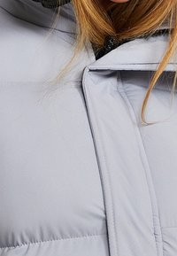 Hellgraue Steppjacke mit hohem Kragen, Reißverschluss und einer weichen, glatten Außenseite mit horizontalen Nähten.