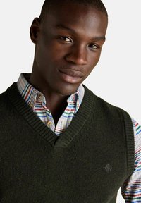 Mladý černoch oblečený v tmavozelenom pletenom svetri cez viacfarebnú pruhovanú košeľu s gombíkmi na jednoliatom svetlom pozadí.