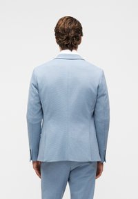 Άνδρας με πλάτη φορώντας σακάκι και παντελόνι σε αν οιχτό μπλε χρώμα μπροστά σε απλό λευκό φόντο.