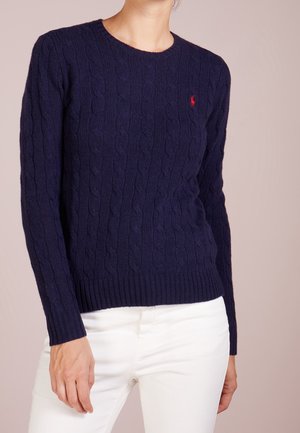 Vrouw draagt een donkerblauwe kabelgebreide trui met een rood logo op de borst en witte broek, staand tegen een effen achtergrond.