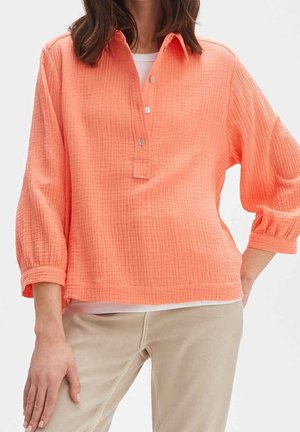 Femme portant une chemise corail ample à manches longues avec des boutons et un pantalon beige, debout avec une main sur la hanche et l'autre détendue.