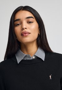 Czarny sweter dzianinowy z okrągłym dekoltem, z białą i czarną kratkowaną kołnierzykiem oraz małym haftowanym logo na piersiach.