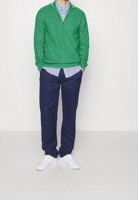 Grön stickad tröja med dragkedja, lagd över en blå och vit randig skjorta. Mörkblå byxor och vita sneakers fulländar outfiten.