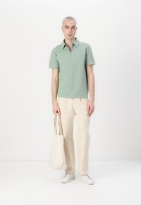 Svetlo zelena polo majica z zadrgo na ovratniku, v kombinaciji s kremnimi hlačami in belimi športnimi copati. Drži svetlo bež torbo.