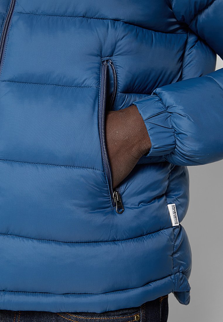 La chaqueta acolchada azul presenta un diseño acolchado, puños elásticos y un bolsillo con cremallera. El material parece brillante y ligero.