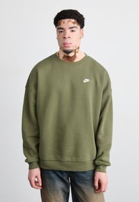 Olivno zeleni Nike sweatshirt s okruglim izrezom i spuštenim ramenskim šavovima. Ima mali bijeli logo na lijevoj strani grudi. Mekana, teksturirana tkanina.