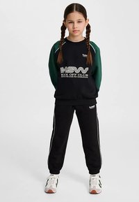 Svart sweatshirt med gröna ärmar, med vit text "Kick Off Club." Matchad med svarta sweatpants med vita detaljer och sneakers.