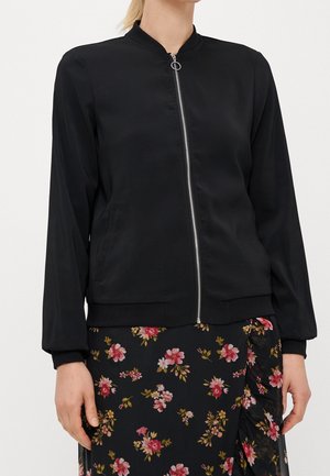 Vrouw draagt een zwarte bomberjack met rits, gecombineerd met een zwarte bloemenrok met roze en rode bloemen.