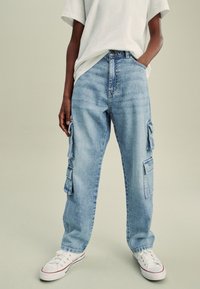 Világoskék cargo farmer, foltzsebekkel, laza szabású és egyenes szárú. Fehér pólóval és fehér sportcipővel viselve.