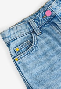 Gros plan de la ceinture d'un jean en denim bleu clair avec poche avant, passants de ceinture, rivets jaunes et bouton rose sur fond blanc.