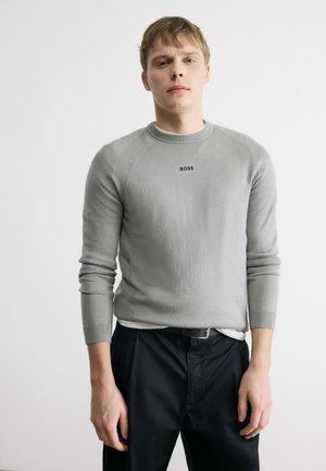 Mladý muž oblečený v svetlosivom svetri s dlhým rukávom s nápisom "BOSS", čiernych nohaviciach a čiernom opasku, stojaci pred bielym pozadím.