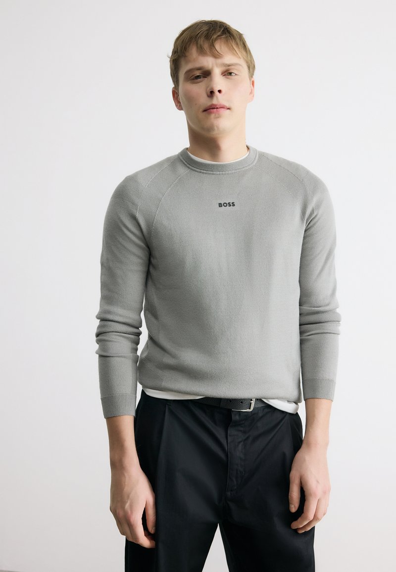 Jeune homme portant un pull léger à manches longues gris clair avec le texte « BOSS », un pantalon noir et une ceinture noire, debout devant un fond blanc uni.