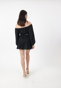 Frau mit langen schwarzen Haaren, die sich umdreht, in einem schwarzen schulterfreien Kleid und weißen High-Heels auf weißem Hintergrund.