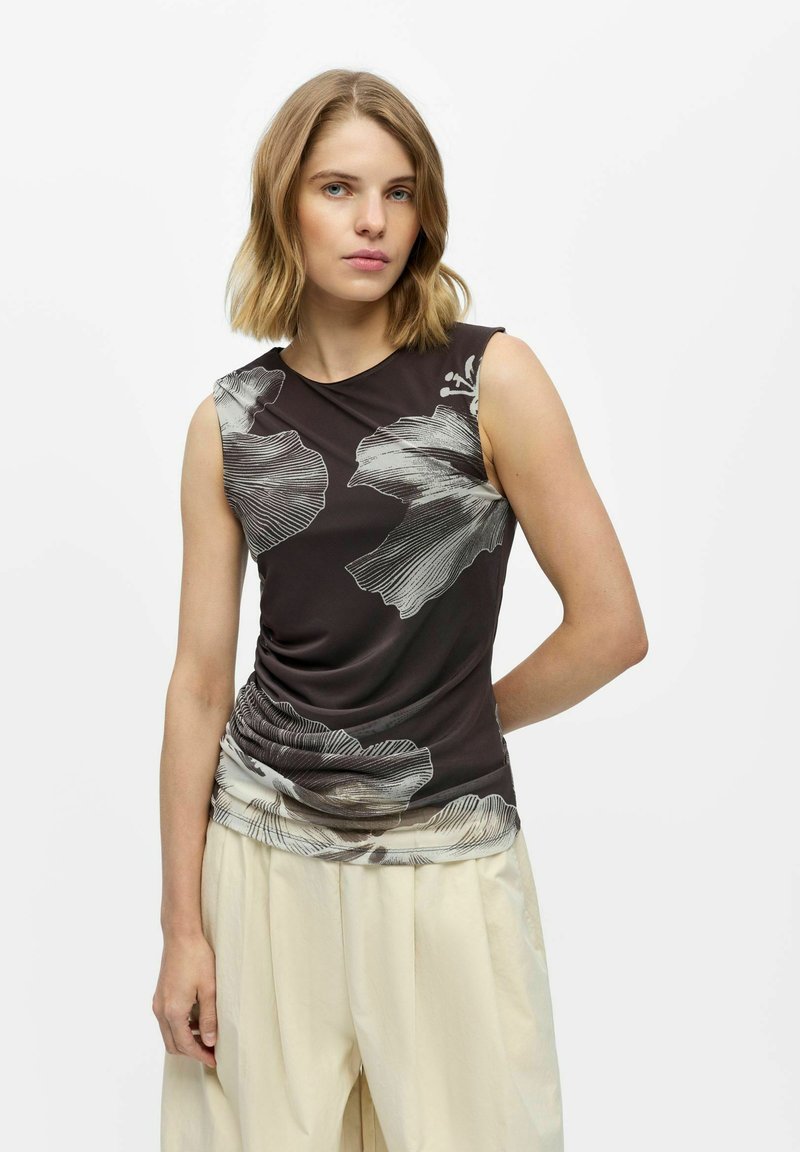 Jeune femme aux cheveux blonds portant un haut noir sans manches avec de grands motifs floraux blancs et un pantalon large beige clair, debout sur un fond blanc.