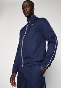 Marineblå træningsjakke med hvide striber, høj krave og lynlås. Har elastiske manchetter og et lille logo på brystet. Glat tekstur.