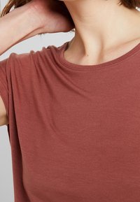 Osoba w rdzawym, krótkim topie, z jedną ręką opartą na szyi, przycięta, aby pokazać szyję i górną część klatki piersiowej.