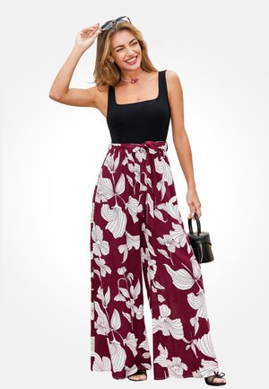 Lächelnde Frau, die ein schwarzes ärmelloses Oberteil trägt, hoch taillierte, weite Blumenhosen in Burgunderrot und Weiß, hält eine Sonnenbrille und eine kleine schwarze Handtasche.