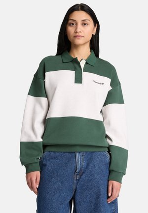 Kvinde iført en grøn- og hvidfarvet langærmet poloshirt og blå jeans, der står mod en ensartet lys baggrund.