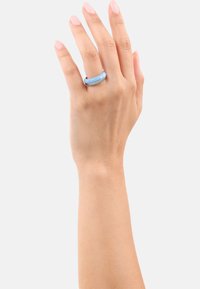 Mano con tono de piel claro que lleva un anillo liso y brillante en azul claro y plata en el dedo medio, sobre un fondo blanco liso.