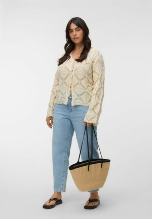 Donna che indossa un cardigan crema con motivo, jeans azzurri chiari, sandali neri, tiene una borsa in paglia beige con manici neri.
