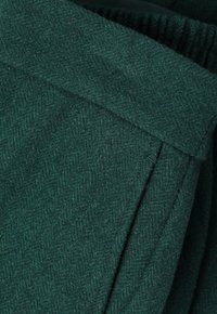 Green herringbone fabric with a soft, textured surface. Features a folded design and visible stitching details, creating a layered look.