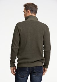 Stickad tröja i mörk olivgrön med ett strukturerat mönster, hög krage och ribbad nederdel. Bärs med mörkblå jeans, sedd bakifrån.