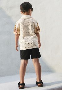 Hämtad topp med beige bas och mönstrade orange ärmar, kombinerad med svarta shorts och mörka sandaler. Sedd bakifrån.