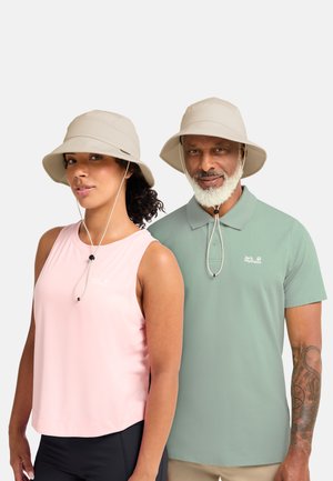 Donna e uomo che indossano cappelli da sole beige con lacci sotto il mento, una canottiera rosa e una polo verde salvia, in piedi davanti a uno sfondo bianco.