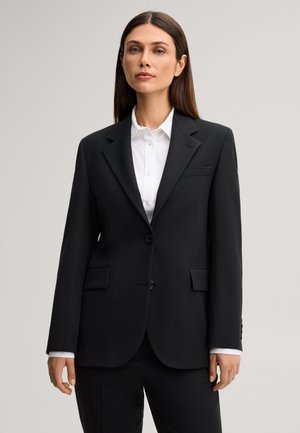 Donna con capelli castani lisci indossa un blazer nero aderente sopra una camicia bianca abbottonata, in piedi davanti a uno sfondo chiaro e uniforme.