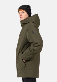 Olivově zelená parka s kapucí, vyrobená z odolného materiálu. Má logo nášivku, boční kapsy a uvolněný střih s hladkou texturou.