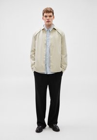 Casaco impermeável bege com botões de pressão, com uma camisa às riscas azuis e brancas por baixo. Calças pretas de perna larga e mocassins.