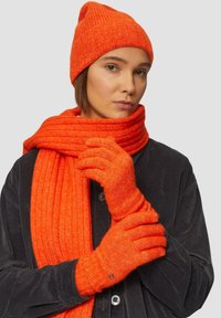 Oranje gebreide beanie, sjaal en handschoenen; geribbelde textuur; effen kleur; handschoenen hebben een cirkelvormig accent. Model draagt een donker overhemd met knopen.
