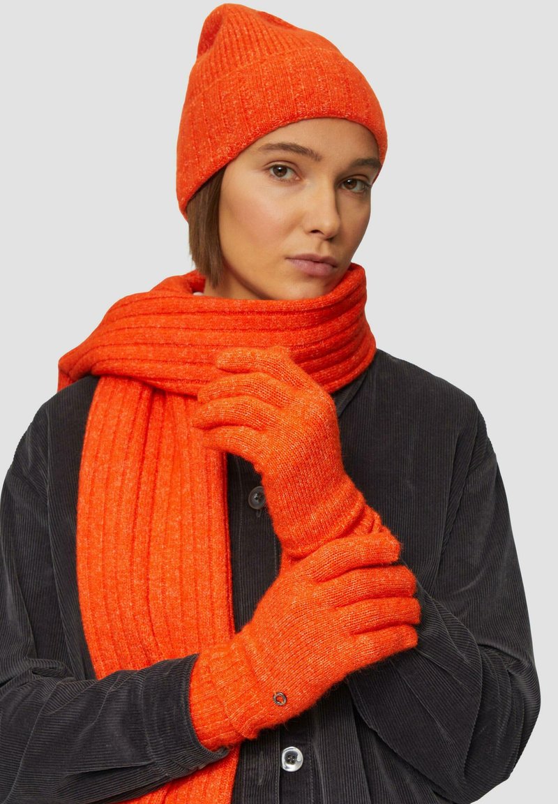 Oranje gebreide beanie, sjaal en handschoenen; geribbelde textuur; effen kleur; handschoenen hebben een cirkelvormig accent. Model draagt een donker overhemd met knopen.