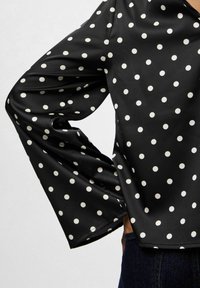 Blusa de satén negra con lunares blancos, mangas largas de campana y un corte holgado. El escote tiene forma de V en la parte delantera.
