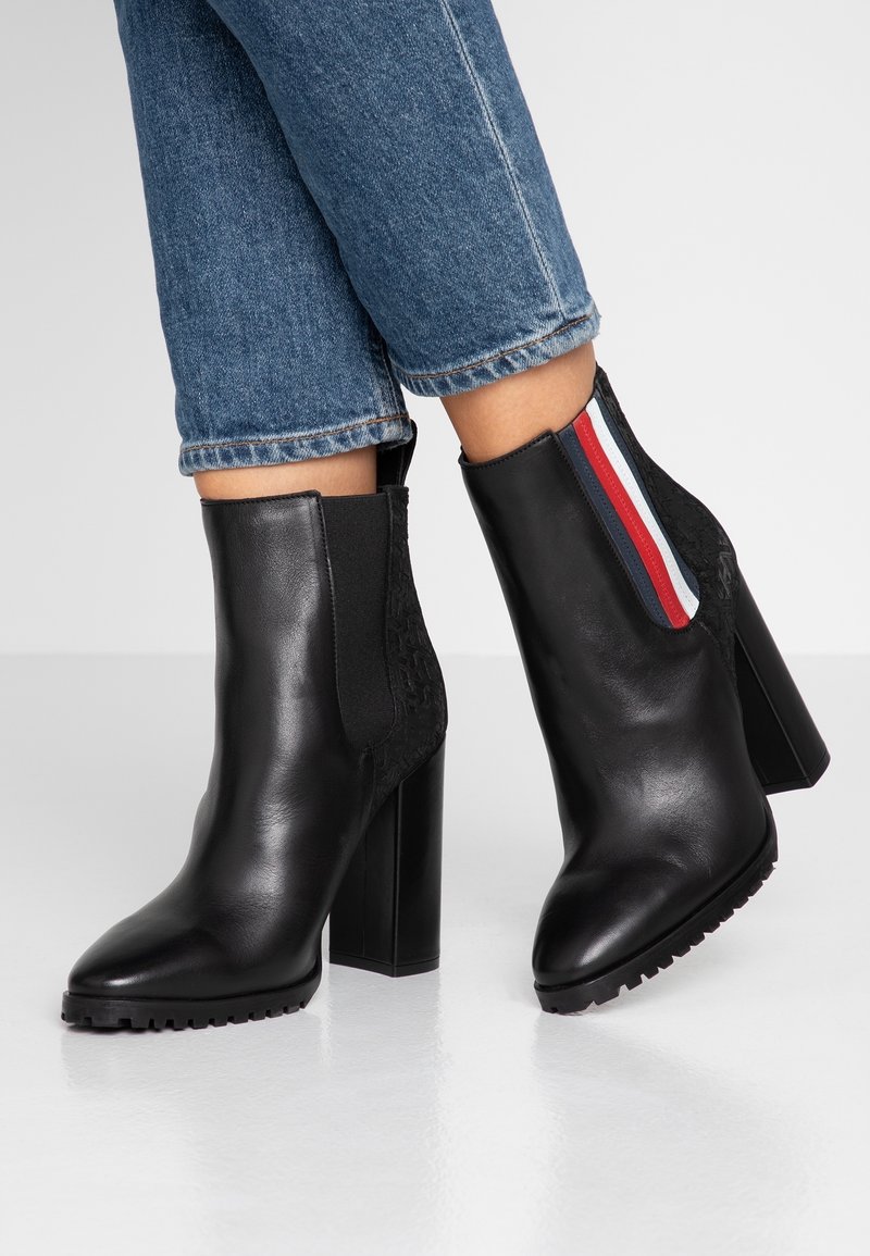 Bottes noires à la cheville avec une finition en cuir verni texturé, talon carré et panneaux latéraux élastiques ornés de rayures rouges, blanches et bleues. Semelle en caoutchouc.